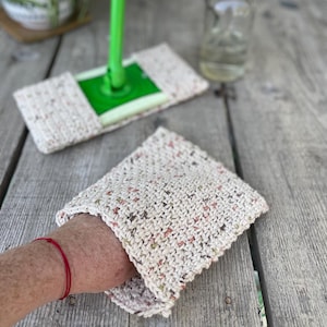 Puede incluir: Primer plano de una cabeza de fregona de crochet con un mango verde y una botella de spray de solución de limpieza. La cabeza de la fregona es beige con motas marrones y rosadas. La botella de spray es transparente y tiene un pulverizador blanco.