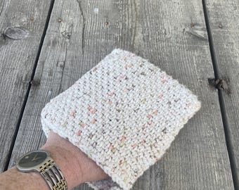 Op de afbeelding: Een witte gebreide ovenwant met roze en groene vlekken. De ovenwant is vierkant en heeft een zacht, getextureerd oppervlak.