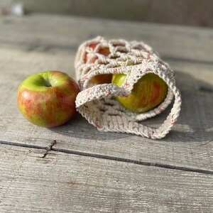 Op de afbeelding: Twee rood-groene appels in een witte gehaakte netzak op een houten oppervlak.