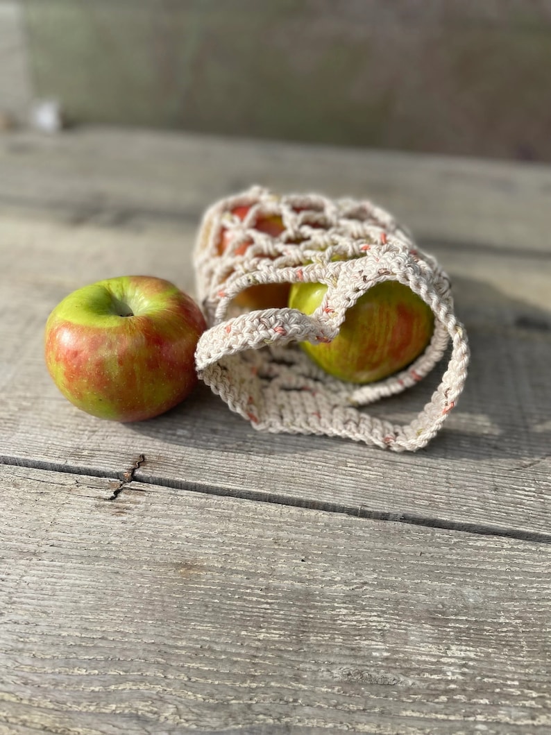 Op de afbeelding: Twee rood-groene appels in een witte gehaakte netzak op een houten oppervlak.