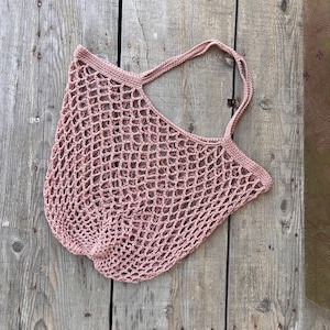 May include: A dusty rose-colored, open-weave net bag with two handles. The bag is made of a knitted material and is sitting on a weathered wooden surface. The bag's design features a large, open mesh pattern.