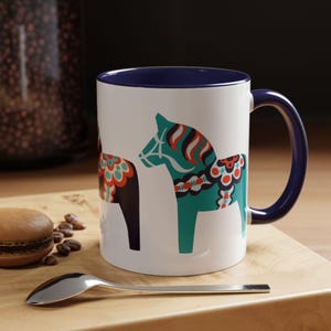 May include: White ceramic mug with a blue rim and handle. The mug features a colorful design of two Swedish Dala horses, one teal and one black, with intricate patterns.