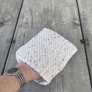 Puede incluir: Un agarrador de olla de punto blanco con motas rosas y verdes. El agarrador de olla es de forma cuadrada y tiene una superficie suave y texturizada.