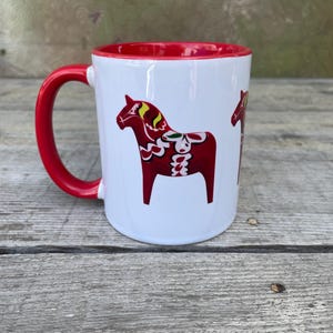 May include: White ceramic mug with a red rim and handle. The mug features a red and white Dala horse design.