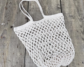 May include: A white, open-weave crochet tote bag with two handles. The bag is made of a light-colored yarn and has a net-like appearance. The bag is empty and sits on a weathered wooden surface.