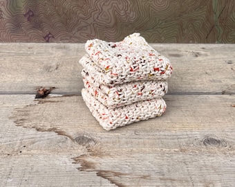 May include: A stack of three square, hand-crocheted dishcloths made from off-white yarn with small, scattered flecks of red, orange, and yellow throughout the woven texture. The cloths are neatly folded and layered on top of one another.