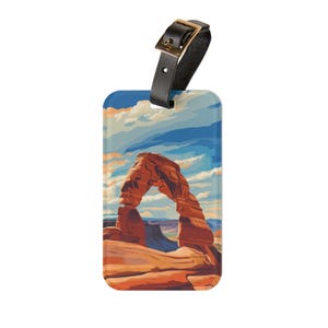 May include: A luggage tag featuring a landscape illustration of a red rock arch under a blue and white sky. The tag has a black leather strap with a gold-colored buckle.