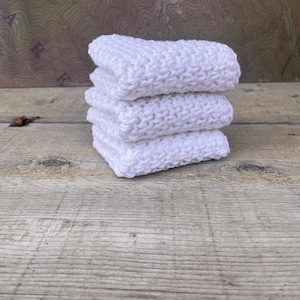 May include: A stack of four white, knitted washcloths. The washcloths are neatly folded and stacked on top of each other. The background is a wooden surface with a map.