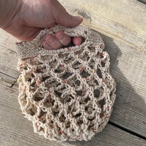 Op de afbeelding: Een gehaakte markt tas met een beige, cr&egrave;me en perzik kleurenschema. De tas heeft een geknoopte handvat en een gaas ontwerp.