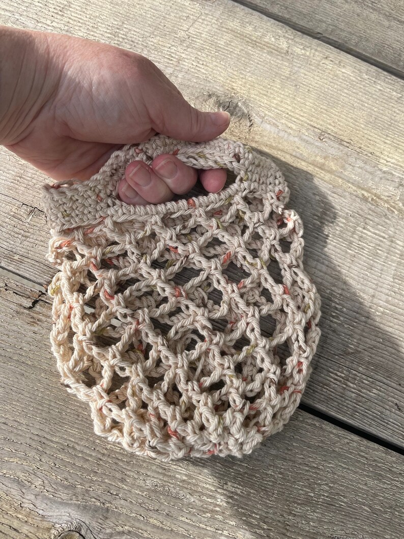 Op de afbeelding: Een gehaakte markt tas met een beige, cr&egrave;me en perzik kleurenschema. De tas heeft een geknoopte handvat en een gaas ontwerp.