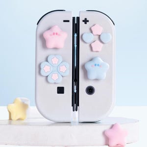 Puede incluir: Un mando de juego gris claro con botones en forma de estrella en rosa pastel, azul y amarillo. El mando tiene un botón más y menos, y un pequeño botón cuadrado negro. El mando está sobre una superficie blanca con un fondo azul claro.