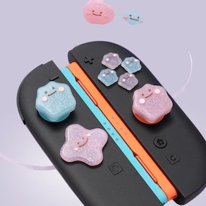May include: A black game controller with orange and blue accents, adorned with several colorful, glittery, cartoon-like thumb grips. The thumb grips are in various pastel colors, including pink, blue, and purple, each with a smiling face.