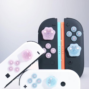 Puede incluir: Mandos Nintendo Switch Joy-Con en blanco y negro, con acentos coloridos. Los mandos presentan botones personalizados y adornos decorativos en forma de pata en rosa y azul, con caras sonrientes.