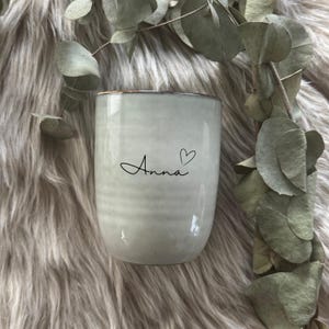 Könnte beinhalten: Eine hellgraue Keramiktasse mit dem Namen "Anna" in Schreibschrift und einem Herzsymbol. Die Tasse hat eine leicht strukturierte Oberfläche und einen braunen Rand. Eukalyptusblätter sind im Hintergrund.