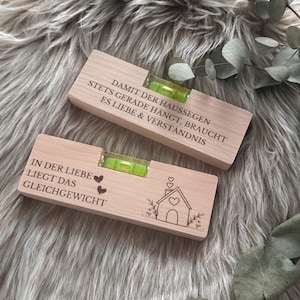 Pode incluir: Dois níveis de madeira com frascos verdes. Um tem o texto "DAMIT DER HAUSSEGEN STETS GERADE HÄNGT, BRAUCHT ES LIEBE & VERSTÄNDNIS". O outro tem o texto "IN DER LIEBE LIEGT DAS GLEICHGEWICHT" com um desenho de uma casa.