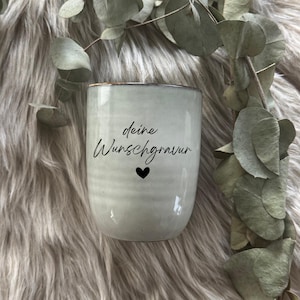 Könnte beinhalten: Eine hellgraue Keramiktasse mit dem Text "deine Wunschgravur" und einem schwarzen Herz. Die Tasse hat eine leicht strukturierte Oberfläche und einen braunen Rand. Eukalyptusblätter sind im Hintergrund.
