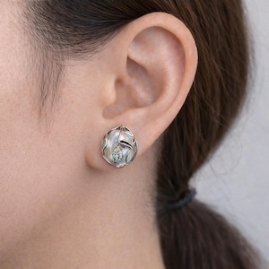 May include: Oval silver earrings with a pearlescent centre and an intricate, openwork design. The earrings have a small, sparkling accent stone and a curved, leaf-like element. The earrings are shown on an ear.