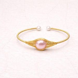 May include: Gold-coloured bracelet with a central pink pearl, framed by a woven gold wire design. Two small white pearls are at the open ends. The jewellery is set against a white background.