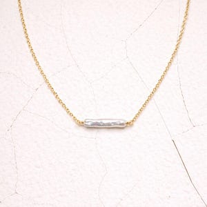 May include: A gold chain necklace with a long, irregular white pearl pendant. The pearl has a textured surface and is suspended horizontally. The necklace is set against a white background.