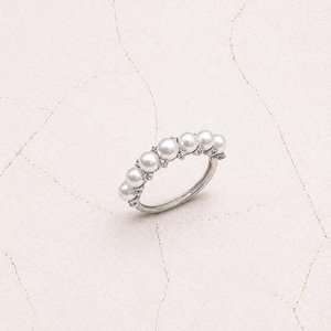 Puede incluir: Un anillo de tono plateado que presenta una hilera de seis perlas blancas redondas dispuestas a lo largo de la parte superior de la banda. Las perlas forman una línea curva con pequeños acentos metálicos entre cada una de ellas.