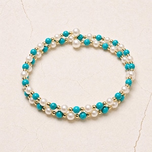 Peut inclure: Un bracelet à double rangée composé de perles blanches, de perles turquoises et d'éléments dorés. Le bracelet est conçu en spirale, créant un effet superposé. Le bijou est présenté sur un fond neutre.