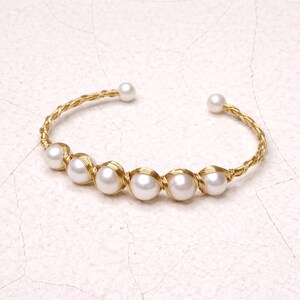 Puede incluir: Brazalete de puño dorado con seis perlas blancas. El brazalete tiene un diseño trenzado y está abierto en los extremos. Las perlas están envueltas individualmente en alambre dorado, creando un aspecto delicado y elegante.