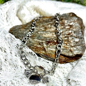 May include: Silver-toned chain bracelet with a variety of charms. Charms include a triangle, a half-circle with embedded crystals, and a leaf. The bracelet is displayed on a textured rock and white lace fabric.