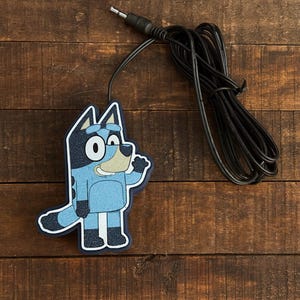 May include: A blue cartoon dog character with a black outline and glitter accents. The character is connected to a black cable with a 3.5mm audio jack. The image is on a brown wooden surface.