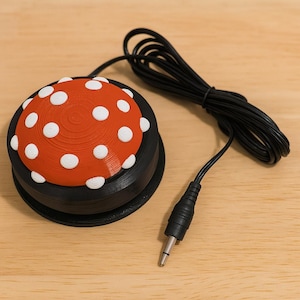 May include: A black and red foot switch with a white polka dot design on the top. The switch has a black cable and a 1/4 inch (6.35 mm) audio jack connector. The switch is on a light brown wooden surface.