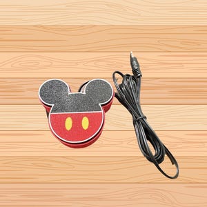 以下が含まれることがあります： 赤と黒のデザインで、黄色の楕円形のアクセントが付いたミッキーマウス型の装飾品。黒いオーディオケーブルがミッキーマウスのアイテムの隣にあります。アイテムは木製の表面にあります。
