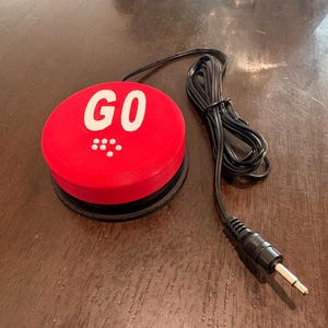 May include: A red button with the word "GO" in white, connected to a black cable and plug. The button has a black base and a series of white dots below the word. The item is on a brown wooden surface.