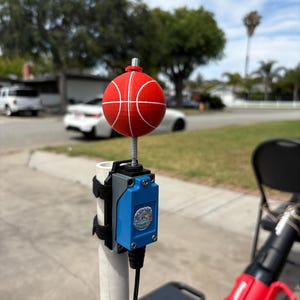 Può includere: Un oggetto rosso a forma di pallone da basket con linee bianche, montato su una molla e un palo di metallo. Una scatola blu e grigia con la scritta "Adaptive Adventure" è attaccata al palo con cinghie nere. Lo sfondo mostra una strada residenziale.