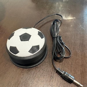 May include: A black and white soccer ball-shaped device with a black base and a black cord with a 3.5mm audio jack. The soccer ball design features white pentagons and black hexagons. The device is likely an audio accessory or a novelty item.
