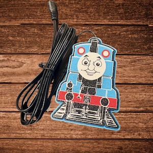 May include: A blue, red, and black train-shaped decorative item with a smiling face, connected to a black cord. The train has a friendly face and is set against a brown wooden background.