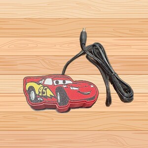 May include: A red Lightning McQueen-shaped night light with a black cord and plug. The car has yellow accents and the number 95. The night light is on a wooden surface.