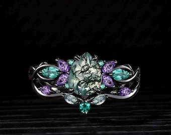 Könnte beinhalten: Ein silberfarbener Ring mit einem großen, ovalen grünen Stein mit komplizierten Mustern. Der Ring ist mit teal- und lilafarbenen Edelsteinen in einem floralen Design verziert. Das Band hat ein detailliertes, verwobenes Muster, das ein elegantes Schmuckstück schafft.