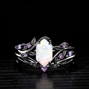 Może przedstawiać: Srebrny pierścionek z dużym, białym, prostokątnym kamieniem centralnym. Pierścionek jest ozdobiony fioletowymi kamieniami akcentującymi i detalami w kształcie liści. Pierścionek jest osadzony na ciemnym tle.