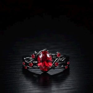 Puede incluir: Un anillo negro con una gran gema roja de corte ovalado en el centro. El anillo presenta un diseño floral con gemas rojas más pequeñas y hojas. El anillo está sobre un fondo oscuro.