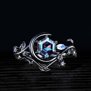 Könnte beinhalten: Silberner Ring mit einem großen, facettierten, sechseckigen Edelstein in Türkis und Lila. Der Ring hat ein Halbmond-Design um den Hauptstein, mit Blatt- und Rankendetails und kleinen Akzentsteinen.