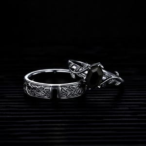 May include: Two silver rings. One ring features a black gemstone and leaf accents. The other ring has a floral design. Both rings are set against a black background.