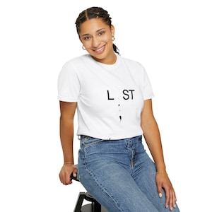 以下が含まれることがあります： 「L ST」の文字と黒いセミコロンのグラフィックがプリントされた白いTシャツ。青いジーンズと組み合わせています。モデルは笑顔で、黒いスツールに座っています。