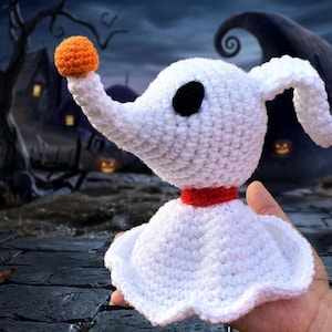 May include: A white crocheted Zero plushie from The Nightmare Before Christmas, featuring a black eye, orange nose, and red collar. The plushie has a ghost-like body and is held in a hand against a spooky background.