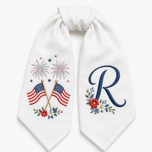 May include: White decorative scarf with embroidered patriotic designs. Features crossed American flags, fireworks, and the letter "R" with floral accents. Perfect for Independence Day celebrations.