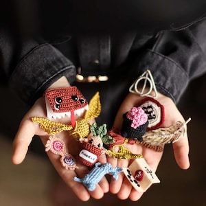 Puede incluir: Una colección de artículos de ganchillo hechos a mano, que incluyen un libro rojo con alas, una snitch dorada, una mandrágora y otros accesorios temáticos. Los artículos son de varios colores, como rojo, dorado, rosa y azul.