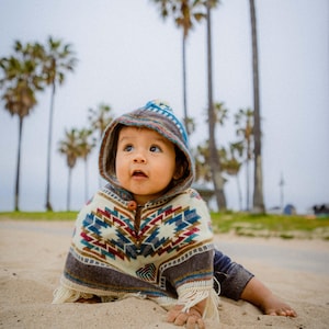 May include: A baby in a hooded poncho with a geometric pattern in blue, red, and cream. The poncho has a fringed hem and a button closure. The baby is on a sandy beach with palm trees in the background.