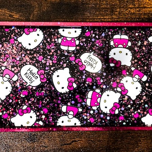 Pode incluir: Uma bandeja retangular com uma moldura rosa e um interior preto brilhante. A bandeja é decorada com personagens Hello Kitty brancos com laços e detalhes rosa. Algumas bolhas de fala dizem "Hello Kitty".