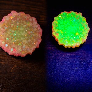 May include: Two resin coasters with a scalloped edge. One coaster is pink and filled with glitter. The other coaster glows bright green. The coasters are on a dark surface and a dark blue surface.