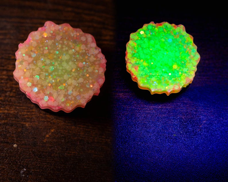 May include: Two resin coasters with a scalloped edge. One coaster is pink and filled with glitter. The other coaster glows bright green. The coasters are on a dark surface and a dark blue surface.