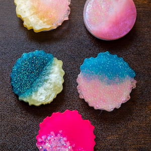 Op de afbeelding: Een assortiment kleurrijke hars coasters. De coasters hebben verschillende ontwerpen, waaronder glitter, verlopen en effen kleuren zoals roze, blauw en geel. De coasters zijn rond met een geschulpte rand.