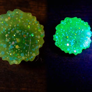 May include: Two round, lime green resin objects with a scalloped edge. The objects are filled with iridescent glitter in various shapes and sizes. One object glows bright green, while the other appears less vibrant.